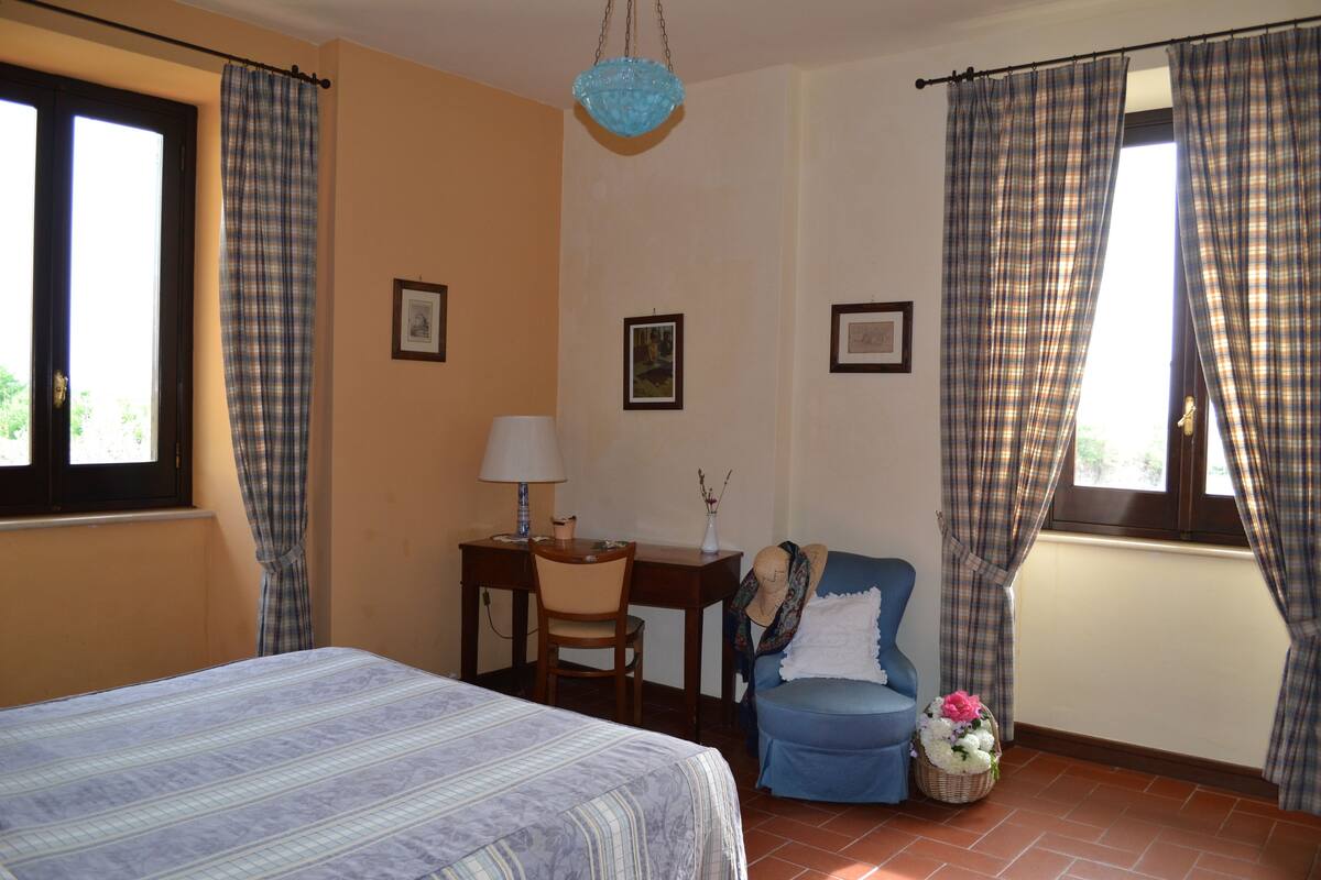 A bedroom with a bed, a desk with a chair, a blue armchair, and a window with curtains.