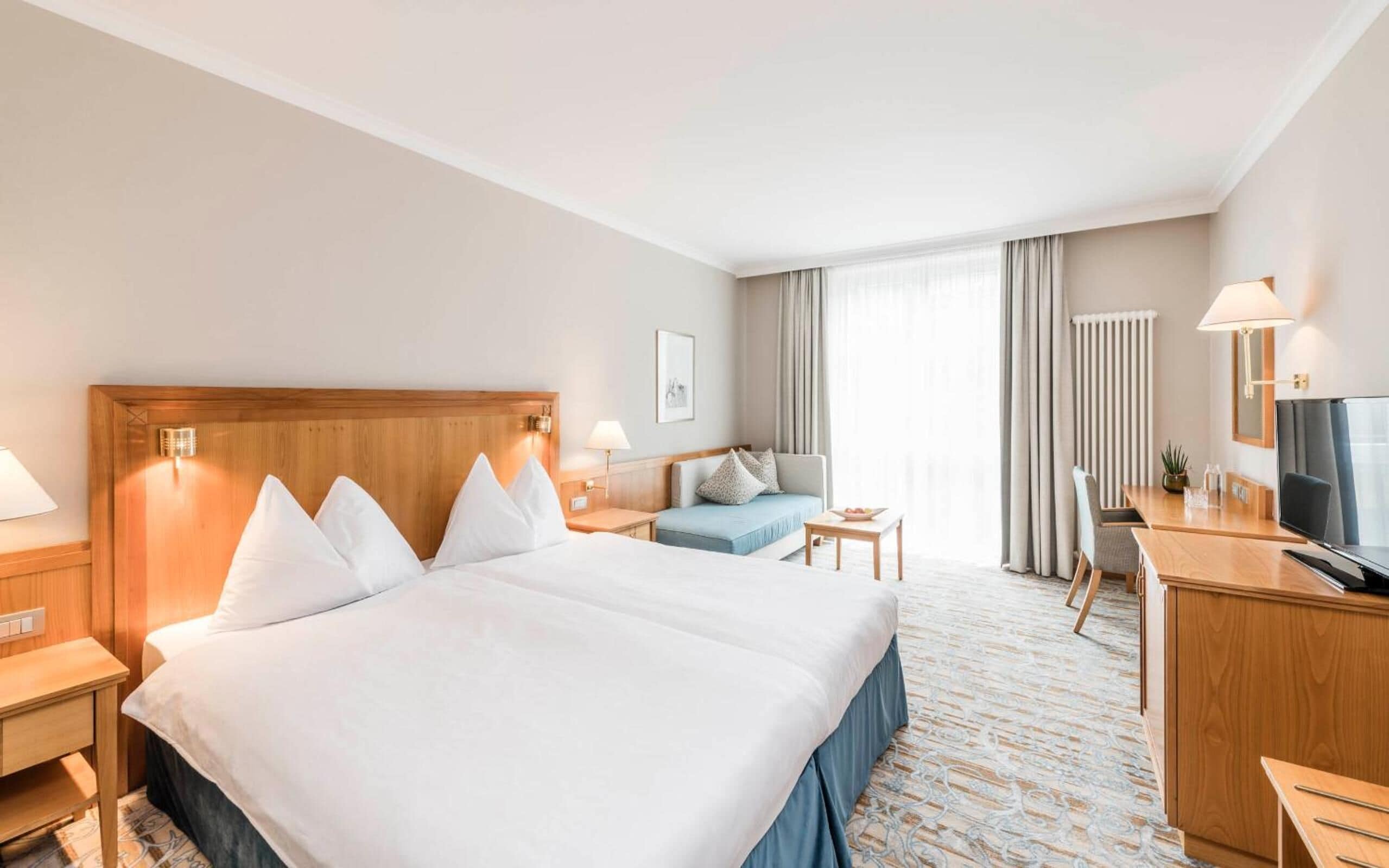 A hotel room with a large bed, a sofa, a desk with a chair, a TV, and a window with curtains.