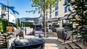 An outdoor seating area with wicker furniture and a wooden deck, overlooking a street with buildings and a restaurant named 'Cosmo'. - Hotel Post (Brunico)