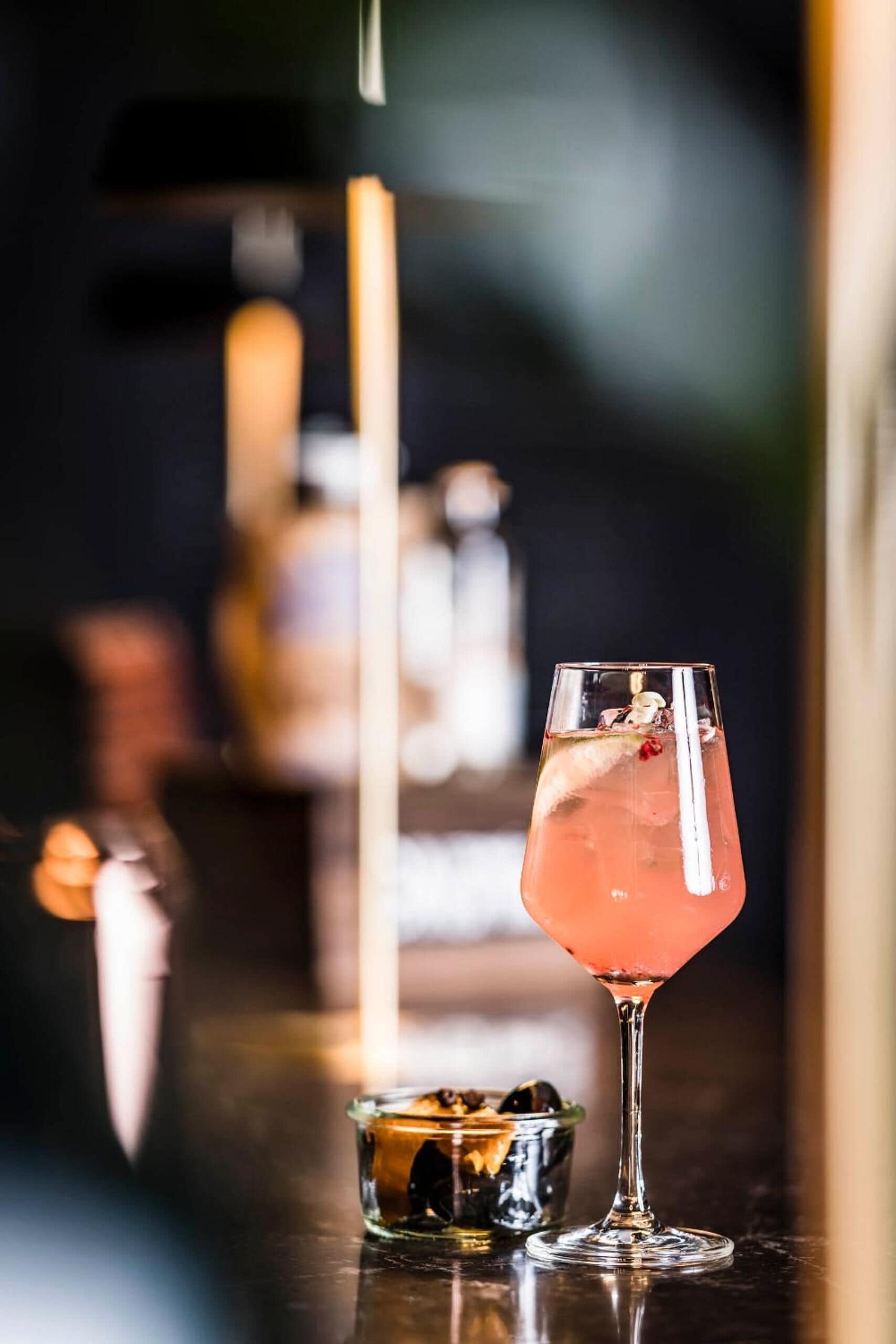 A cocktail glass with a pink drink and a small bowl of ice cubes on a bar counter.