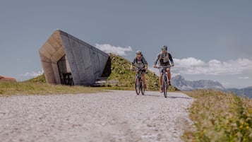 Dos ciclistas en un camino de grava con un edificio moderno y montañas al fondo.