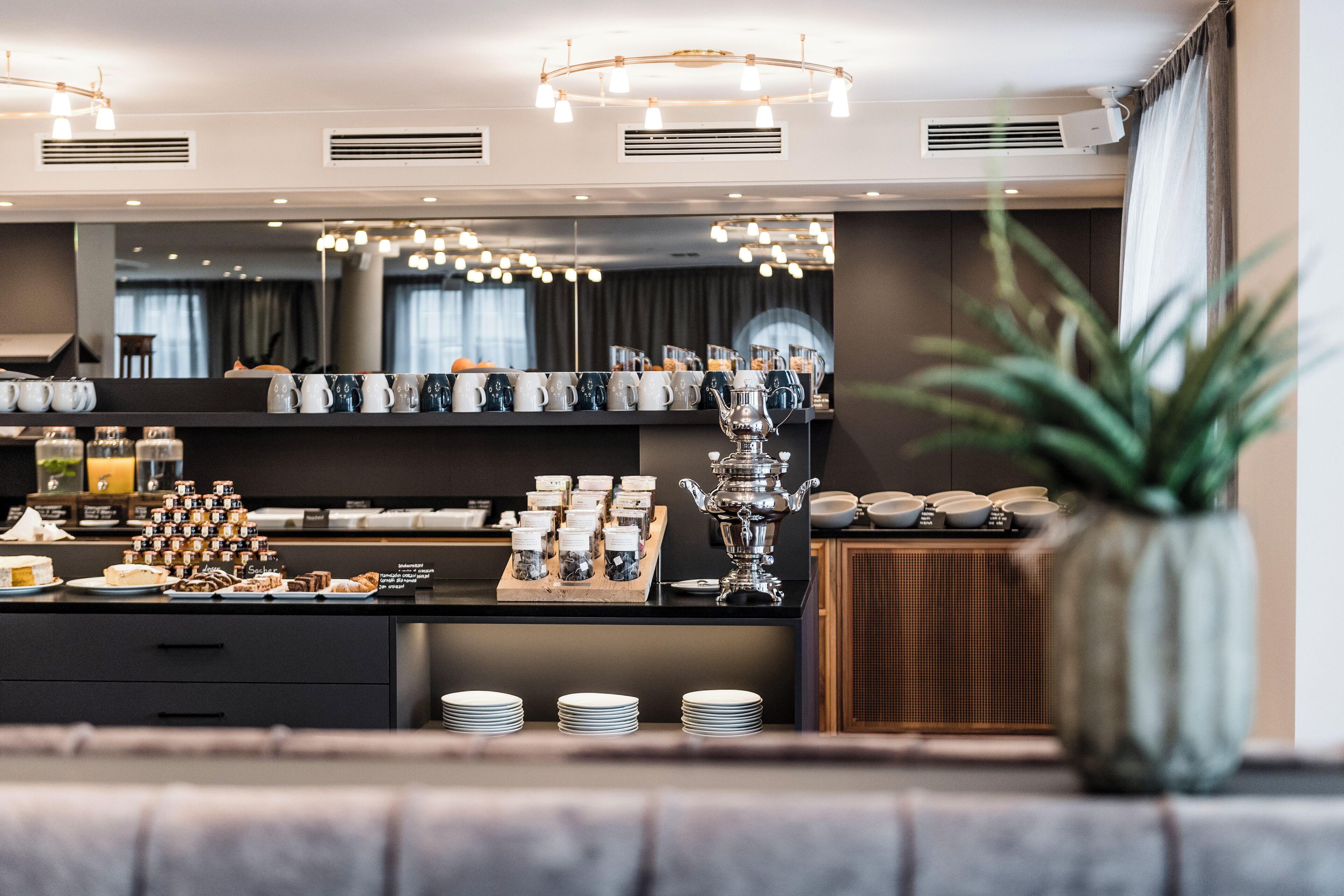 A modern hotel breakfast area with a buffet setup, including various food items and a decorative silver urn.