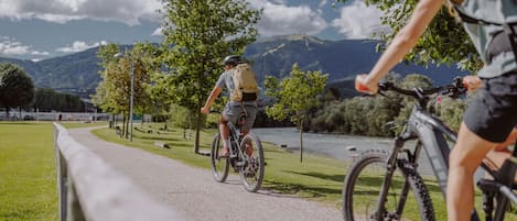 A paved path with cyclists, surrounded by greenery and a lake, with mountains in the background.