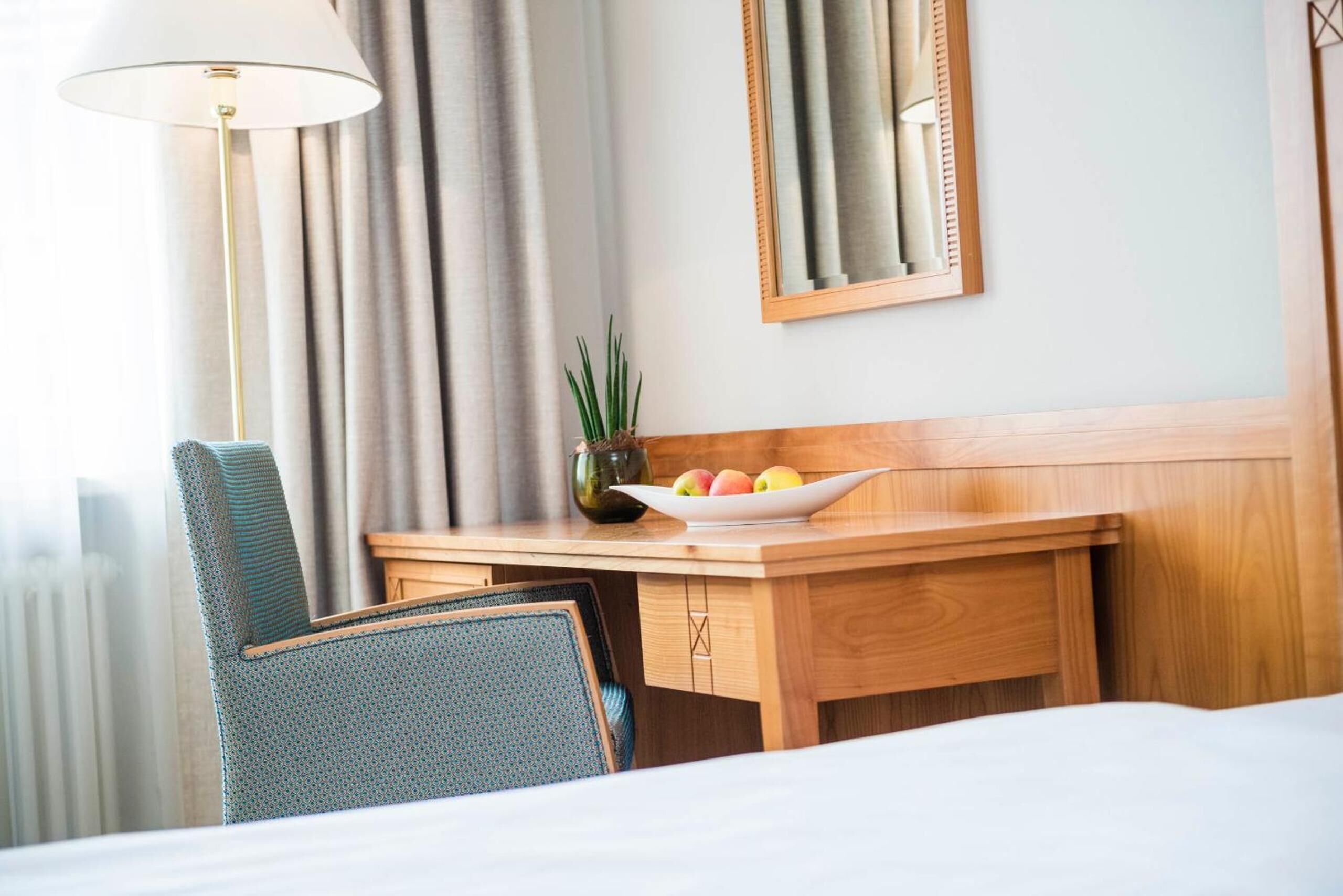A hotel room with a wooden desk, a bowl of fruit, a potted plant, a lamp, and a mirror on the wall.
