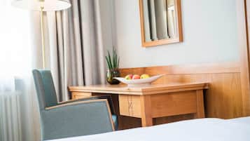 A hotel room with a wooden desk, a bowl of fruit, a potted plant, a lamp, and a mirror on the wall.