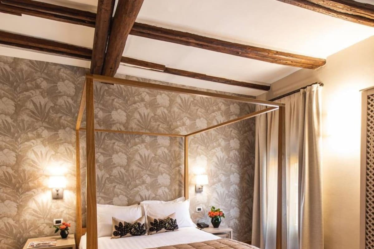 Een hemelbed met houten frame, bloemen behang en een nachtkastje met een lamp.
