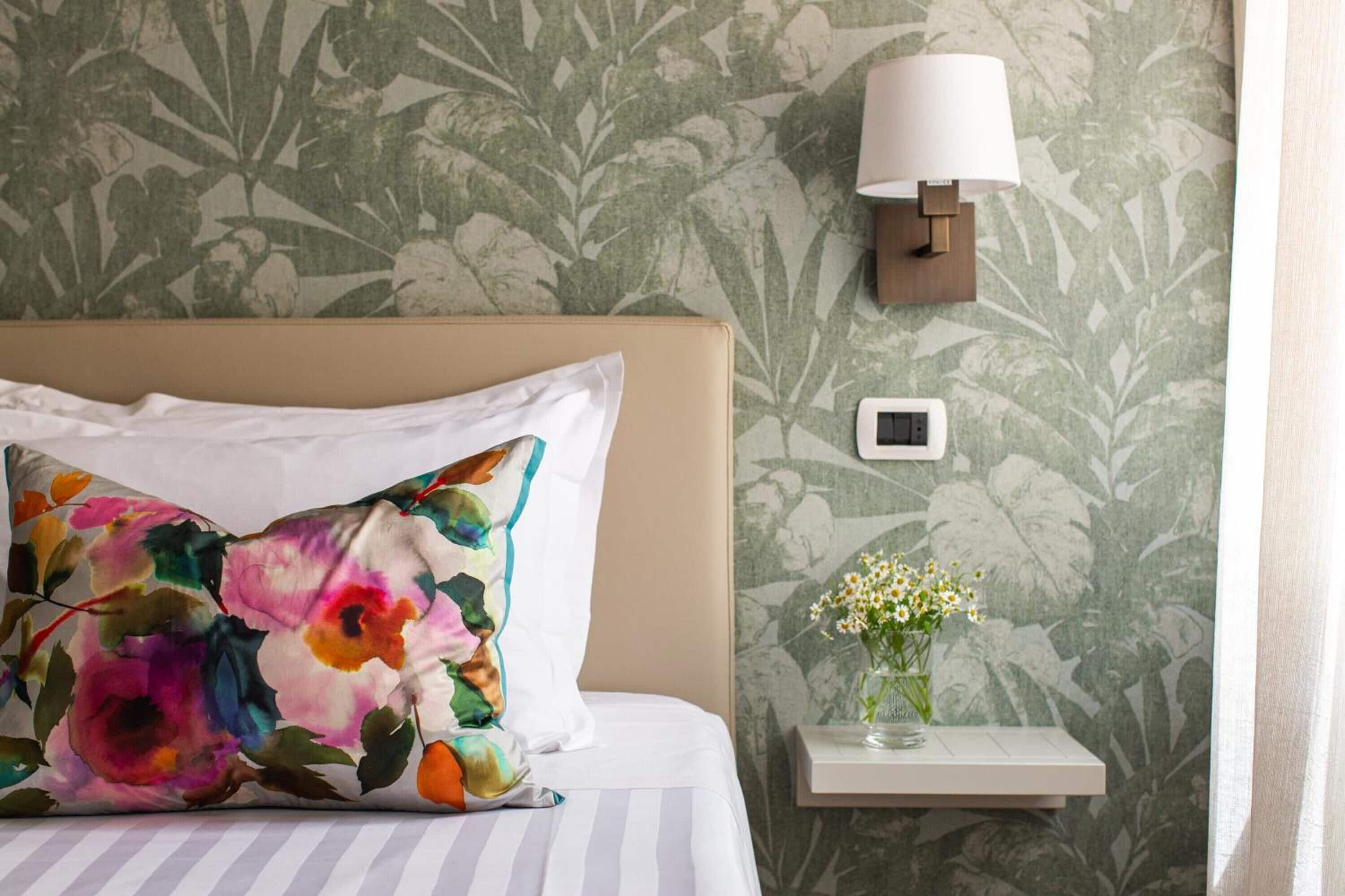 A hotel room with a bed, floral-patterned wallpaper, a bedside lamp, a small table with a vase of flowers, and a thermostat on the wall.
