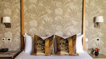 Habitación de hotel con cama, papel tapiz floral, cabecera de madera, lámparas de noche y mesita de noche con un jarrón de flores.