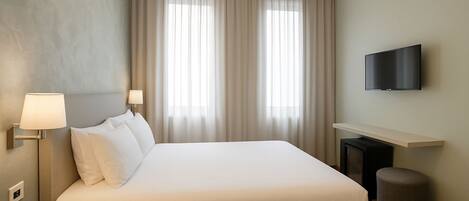Habitación de hotel con una cama grande, un televisor de pantalla plana, una mesita de noche con lámpara y un otoman redondo pequeño.