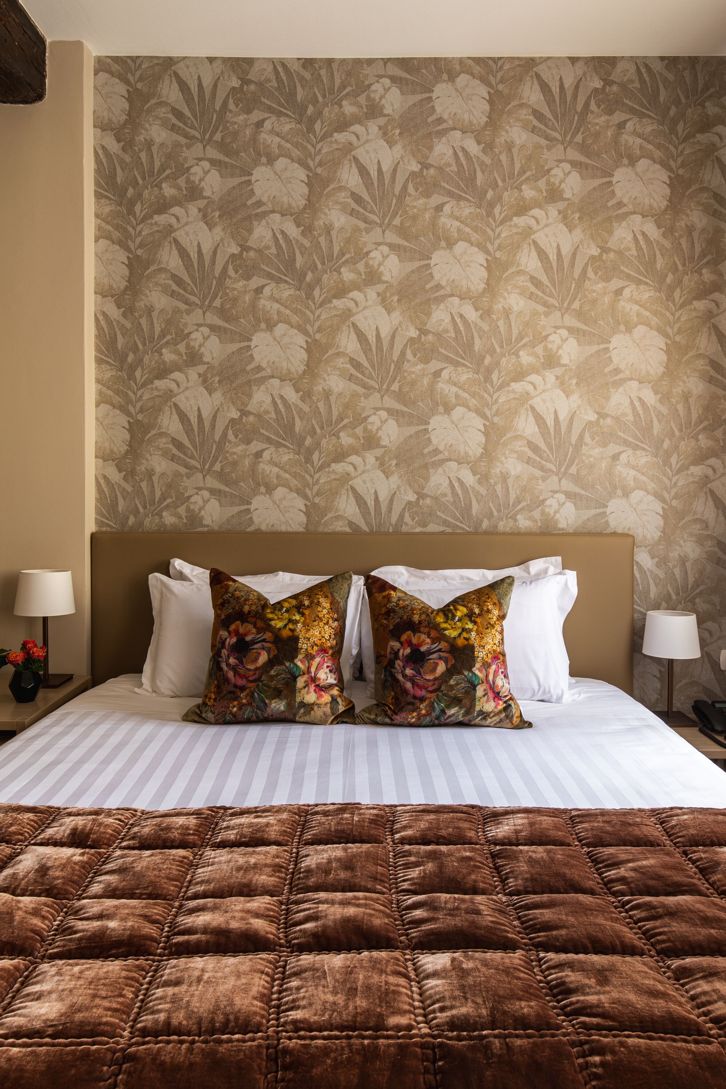 A neatly made bed with a brown quilt, two floral pillows, and a headboard against a floral wallpaper background.