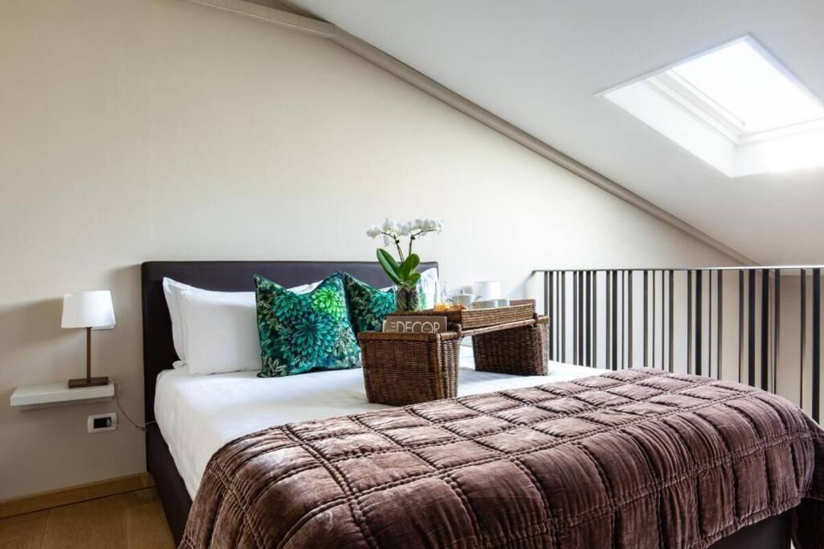 Un lit bien fait, recouvert d’une couverture marron, avec un oreiller blanc et un oreiller vert. Une petite table avec une lampe se trouve à côté du lit. Un panier en osier contenant une plante est posé sur le lit.
