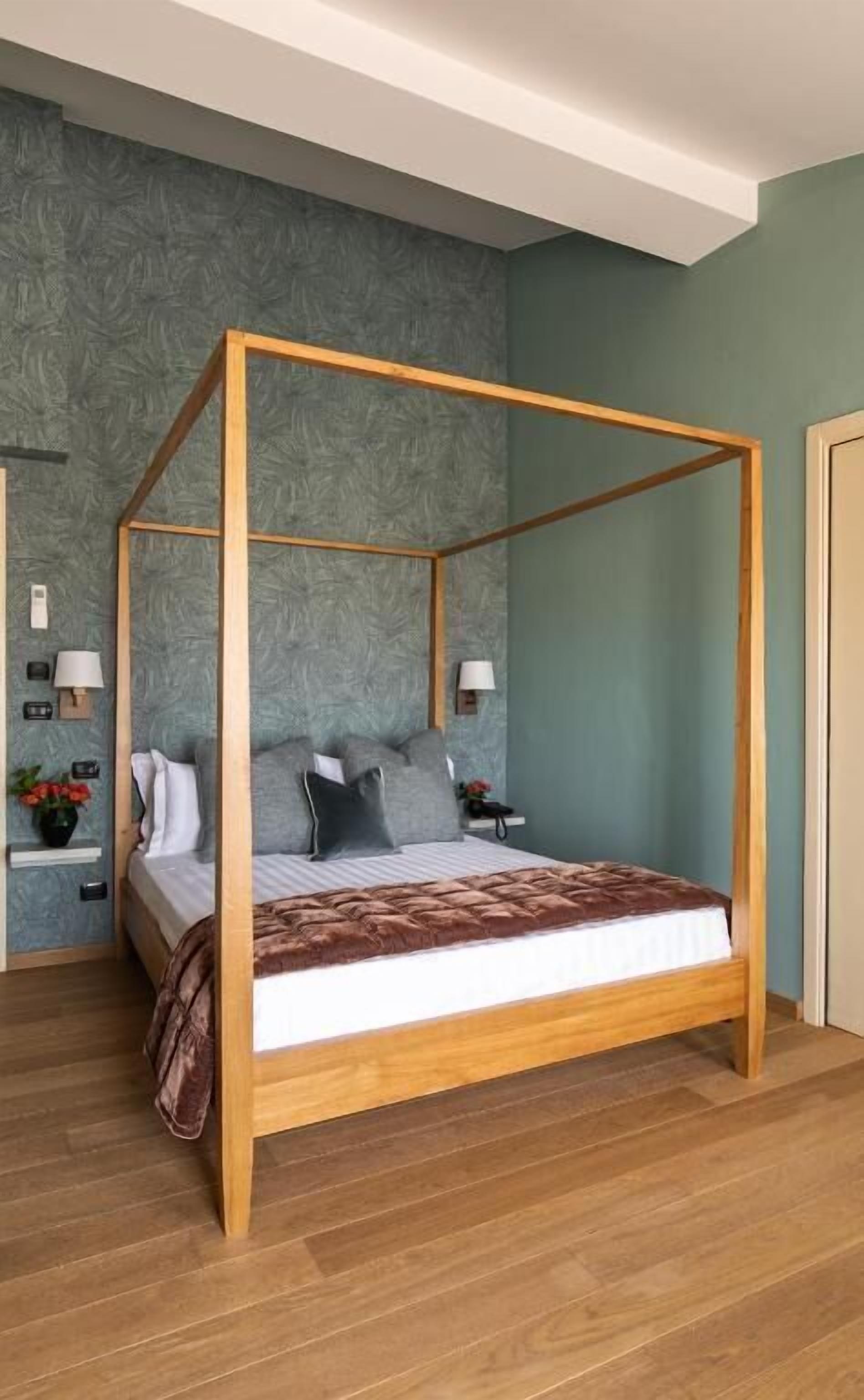 Ein Bett mit Himmel und Holzrahmen, ein grauer Stoffkopfteil und eine braune Tagesdecke. Auf einem der beiden Nachttische stehen eine Lampe und eine kleine Pflanze. Der Raum hat Holzböden und eine Wand mit strukturierter Grau-Oberfläche.