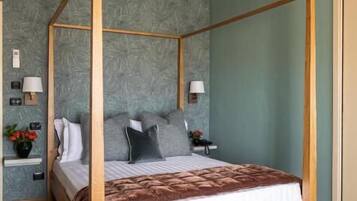 A four-poster bed with a wooden frame, a grey upholstered headboard, and a brown throw blanket. There are two bedside tables with lamps and a small plant on one. The room has wooden flooring and a textured grey wall.