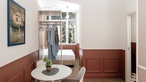 A room with a bed, a closet with hanging clothes, a table with a chair, and a window with a view of buildings. - Corso Italia Suites (Sorrento)