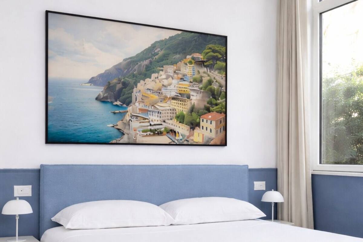 A bedroom with a bed, a nightstand, a lamp, a window, and a painting of a coastal town.