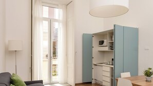 A modern living room with a grey sofa, green cushions, a dining table with chairs, a kitchen area with cabinets, and a large white pendant light. - Corso Italia Suites (Sorrento)