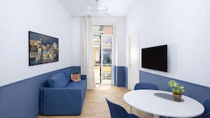 A modern room with a blue sofa, a round table with chairs, a TV mounted on the wall, and a painting of a coastal town. - Corso Italia Suites (Sorrento)