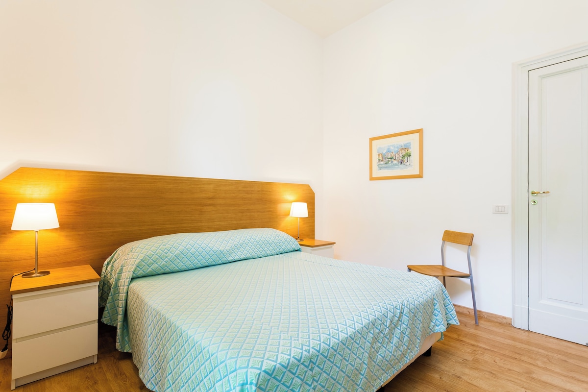 Una cama bien hecha con una colcha estampada, dos lámparas de noche, una mesita de noche de madera, una silla y un cuadro en la pared.