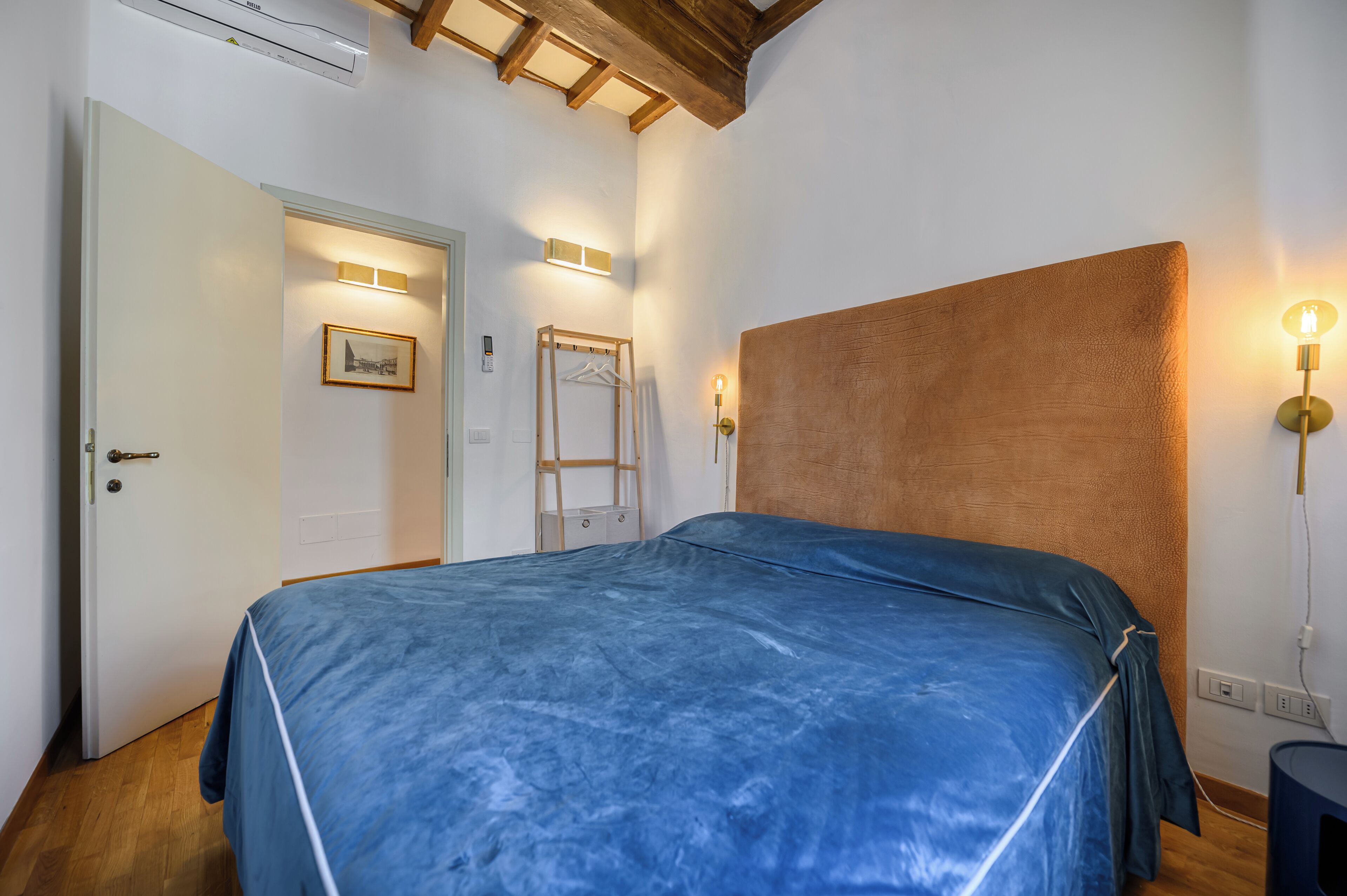 A bedroom with a large bed, wooden ceiling beams, a white door, and a wall-mounted light fixture.
