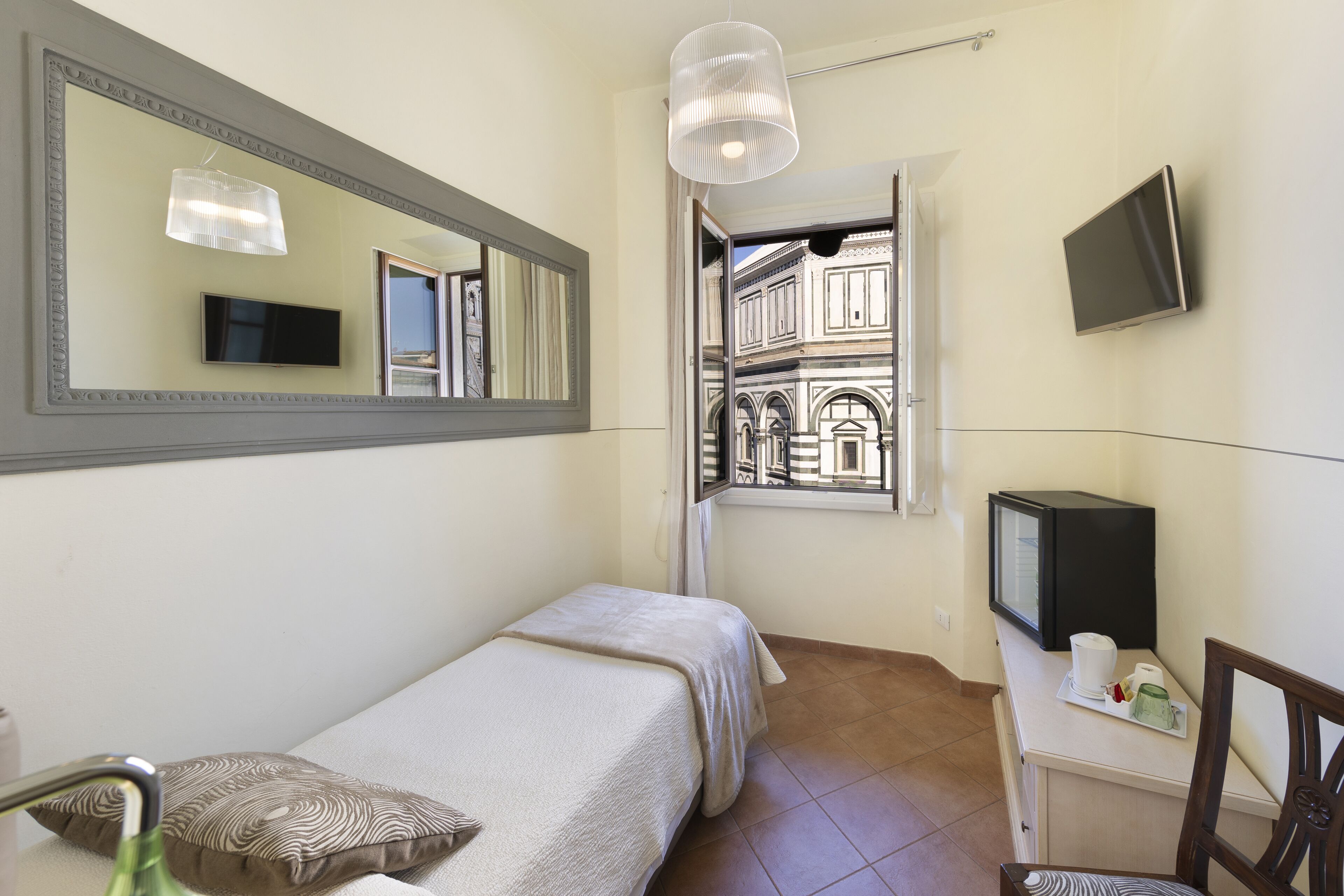 A bedroom with a bed, a TV, a mirror, and a window with a view of buildings.