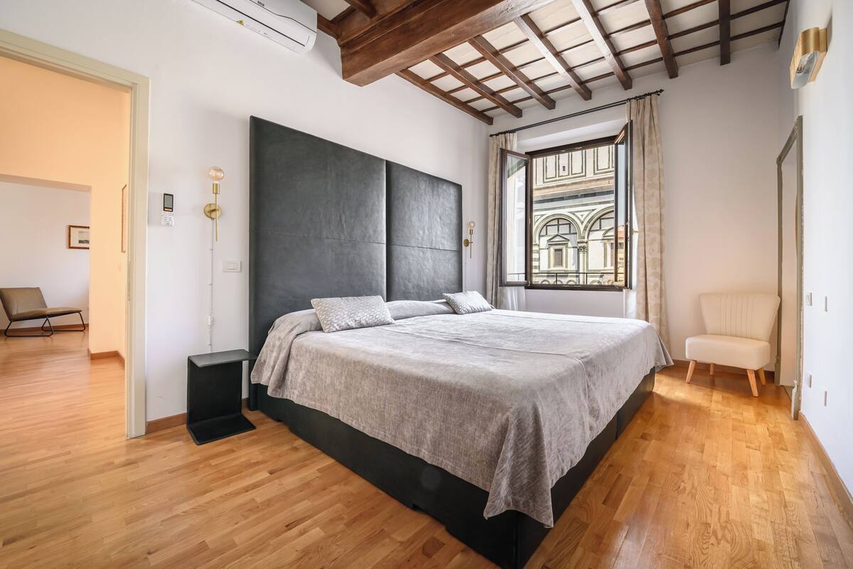 A modern bedroom with a large bed, a built-in bench, a view of a courtyard through a window, and a wooden floor.