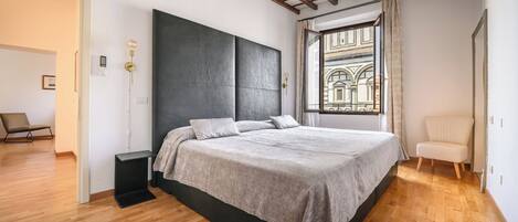 A modern bedroom with a large bed, a built-in bench, a view of a courtyard through a window, and a wooden floor.