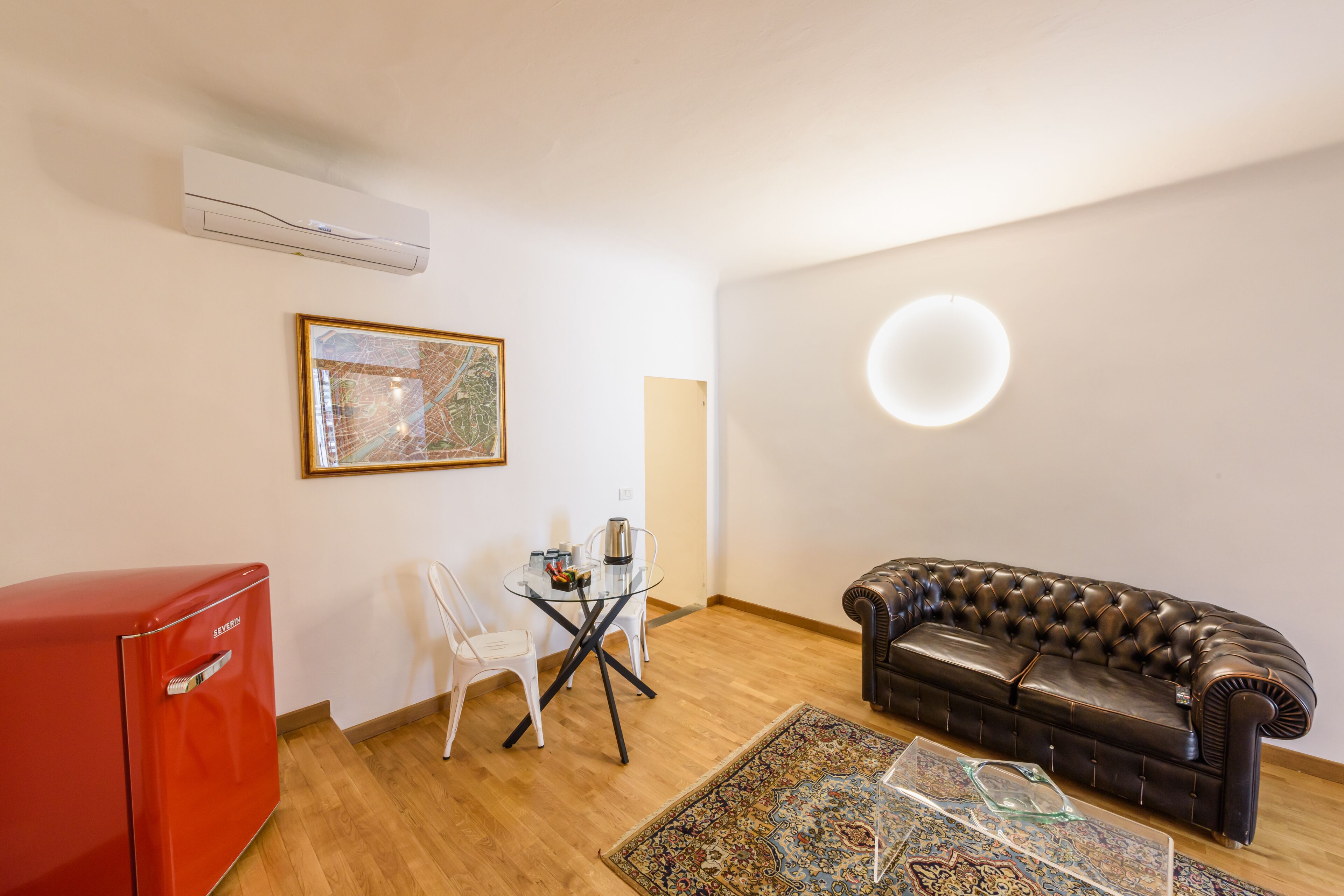 A compact living space with a red refrigerator, a glass table with chairs, a black leather sofa, a patterned rug, and a wall-mounted air conditioner.