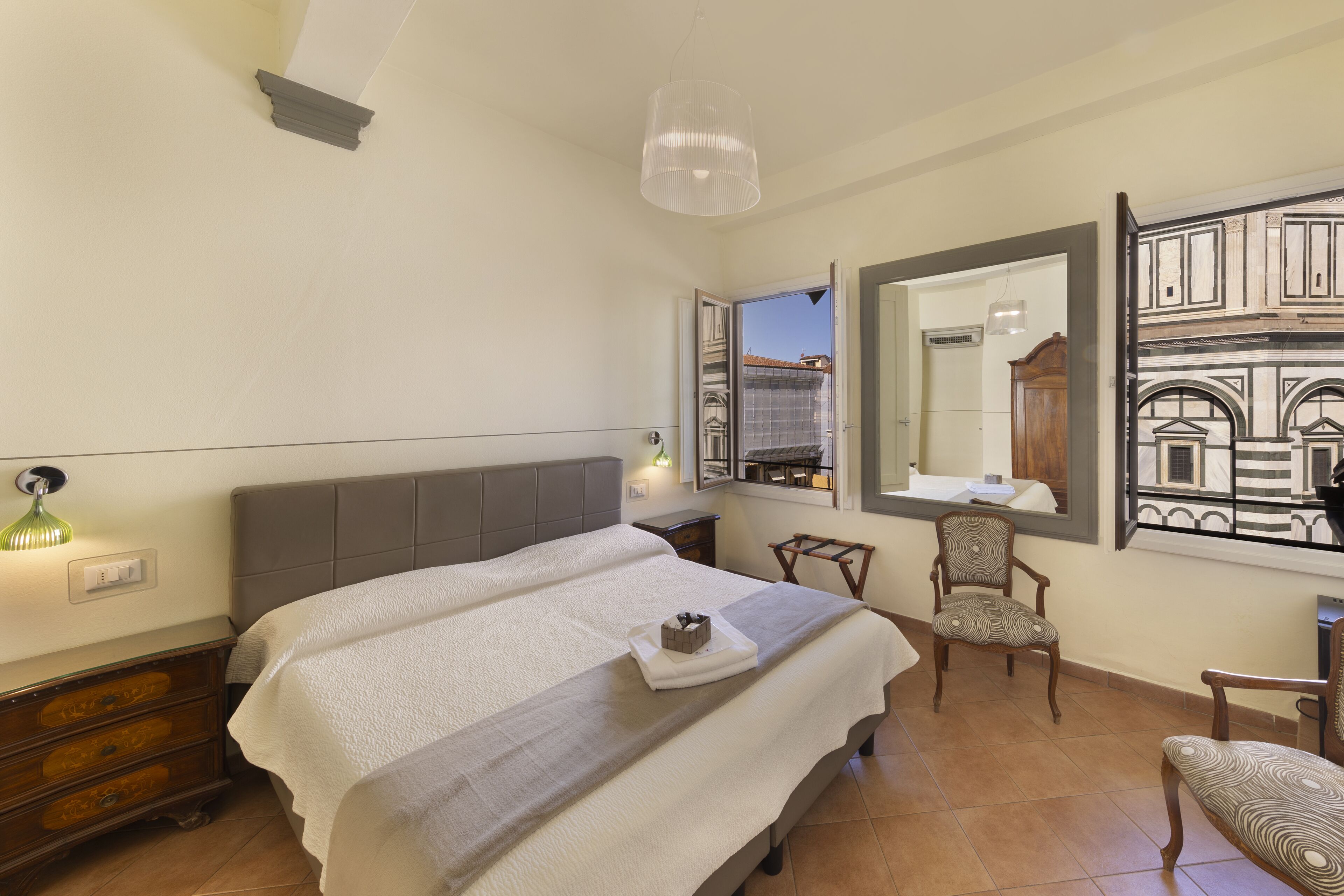 A bedroom with a bed, a chair, a desk, and a large mirror reflecting a building.