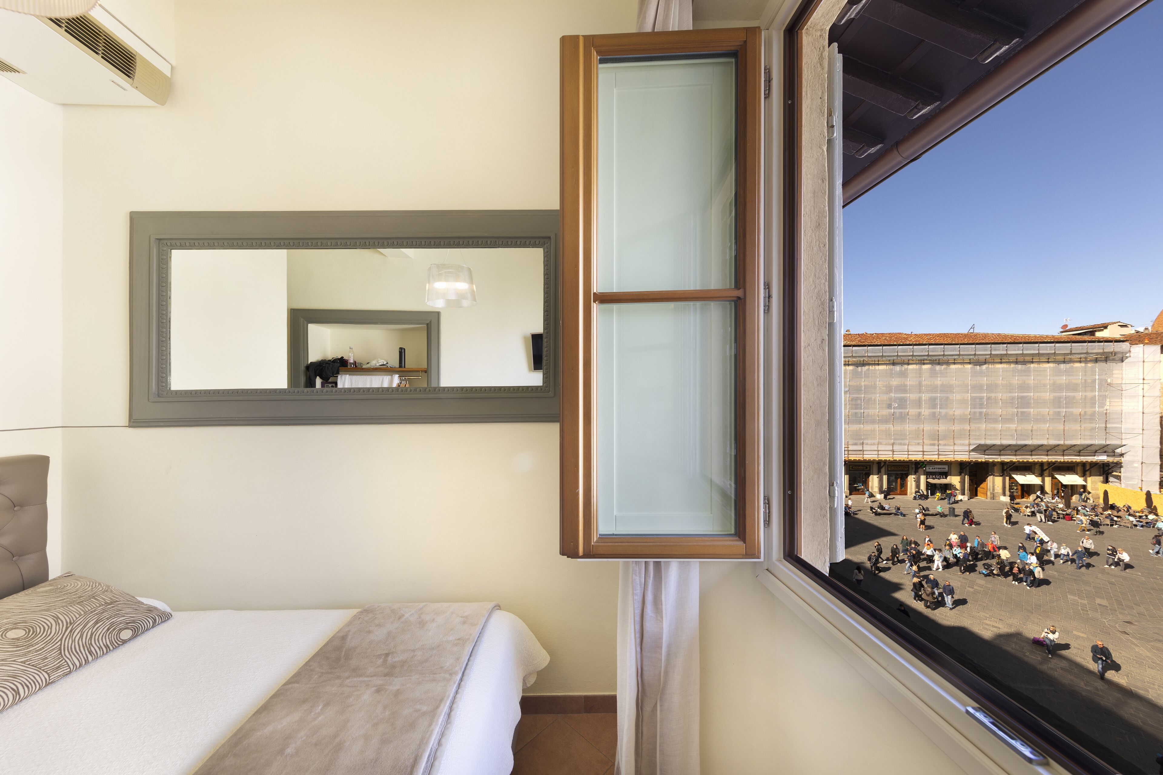 A hotel room with a bed, a large mirror, and a window overlooking a plaza.