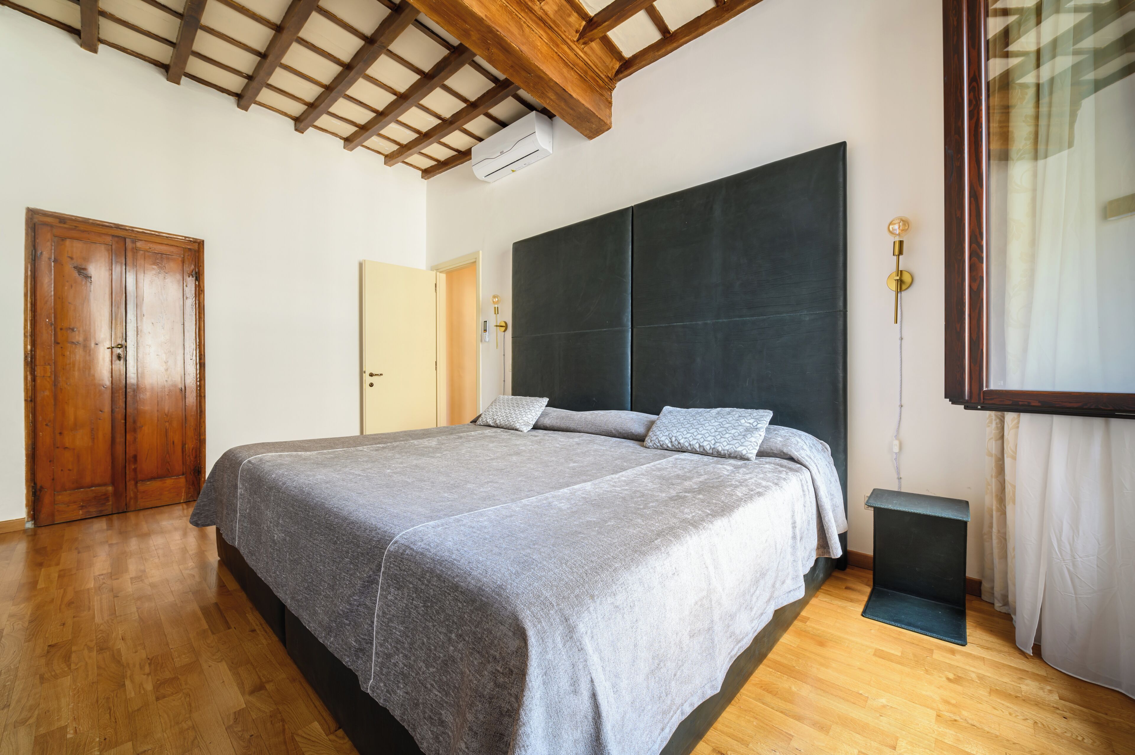 A bedroom with a large bed, wooden flooring, a dark headboard, a small bedside table, and a wooden door.