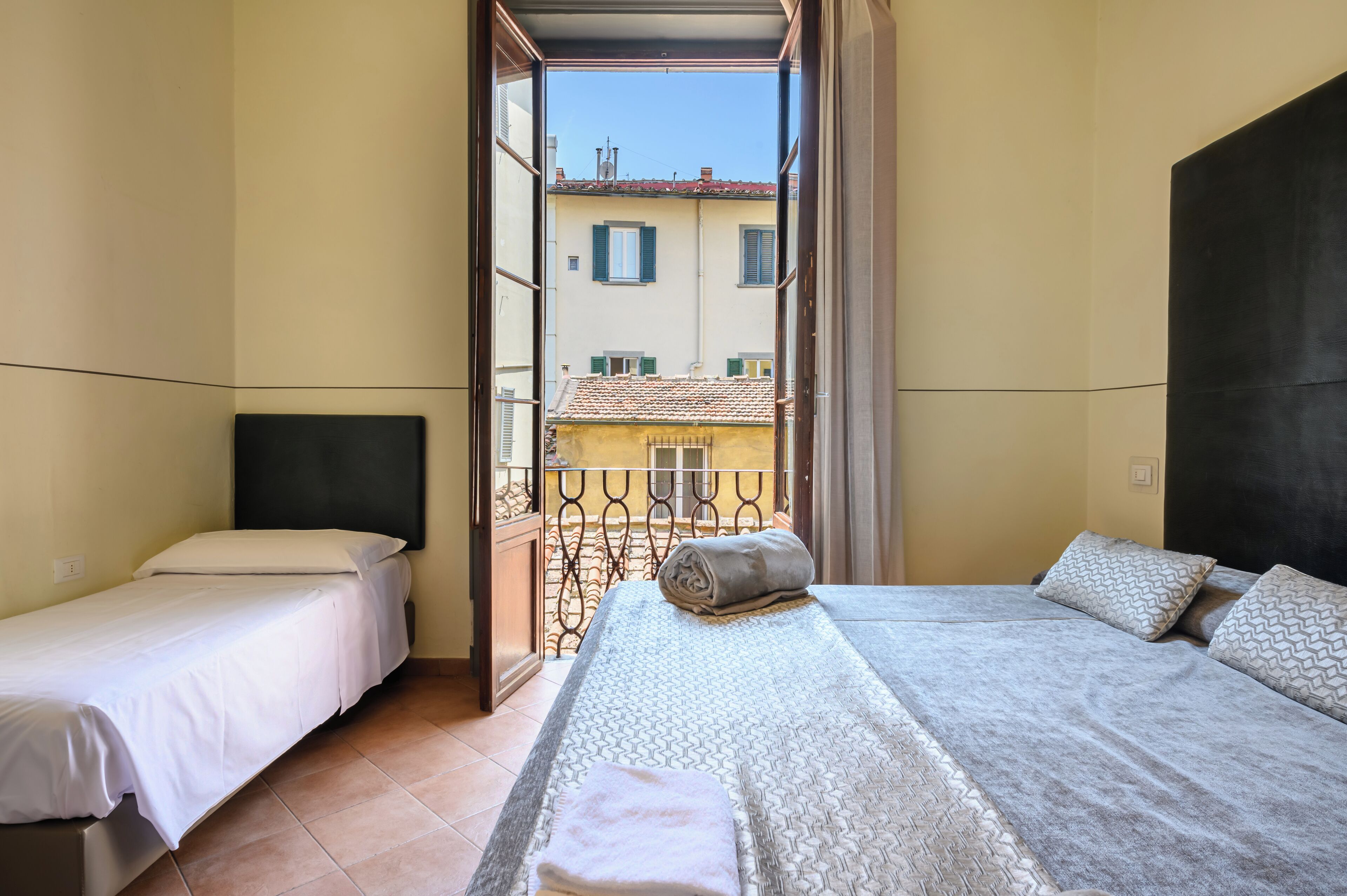 A room with two beds, a balcony with a metal railing, and a view of buildings in the background.