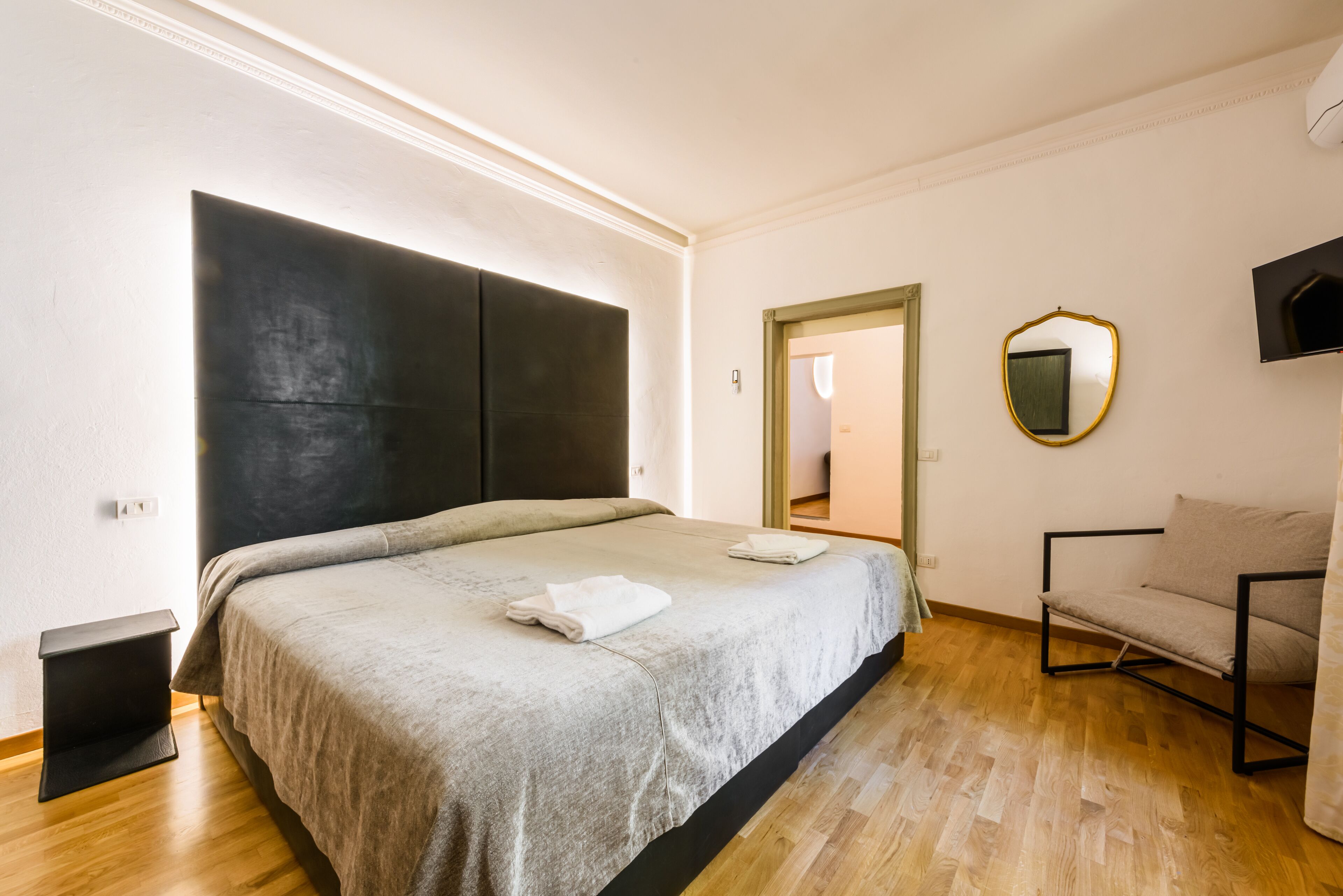 A modern bedroom with a large bed, a small bedside table, a chair, and a mirror on the wall.