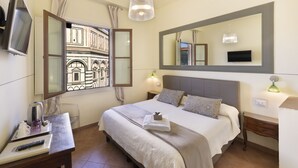 A hotel room with a bed, a desk, a TV, a window with a view of a building, and a mirror on the wall. - Soggiorno Battistero (Florence)