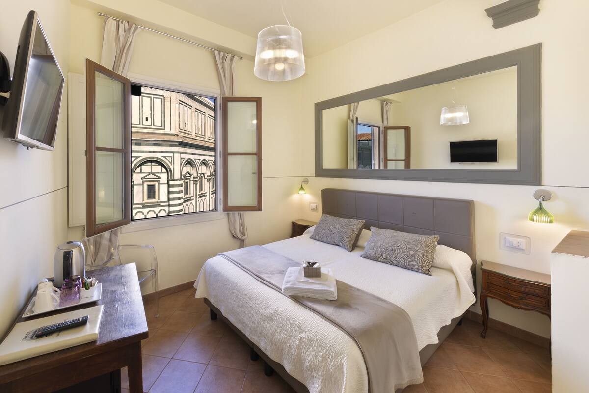 A hotel room with a bed, a desk, a TV, a window with a view of a building, and a mirror on the wall.