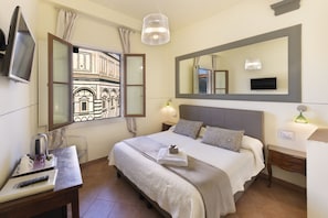 Habitación de hotel con cama, escritorio, televisor, ventana con vista a un edificio y espejo en la pared.