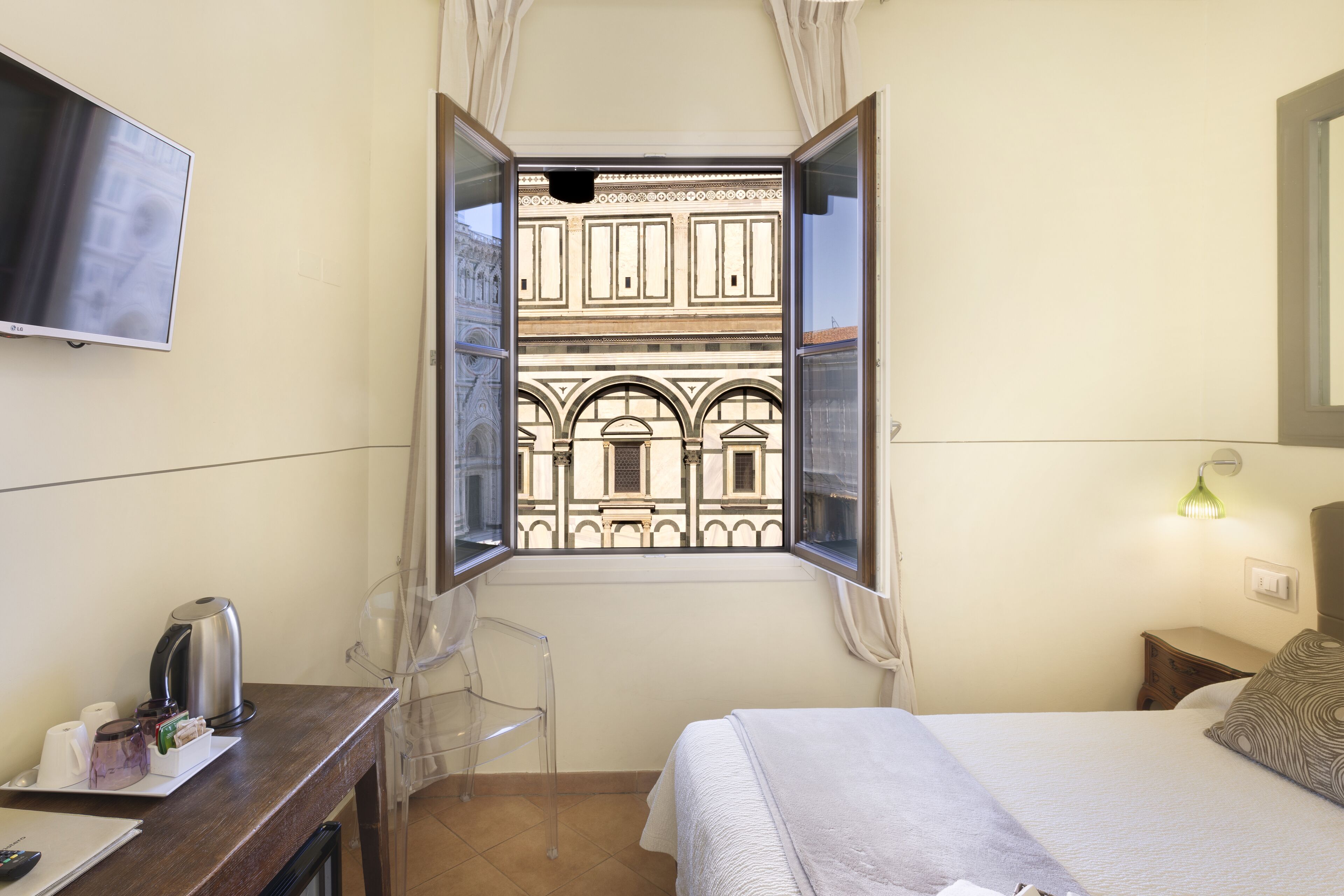 Ein Hotelzimmer mit einem Bett, einem Schreibtisch mit Wasserkocher, einem Sessel, einem Fernseher und einem Fenster mit Blick auf ein Gebäude.