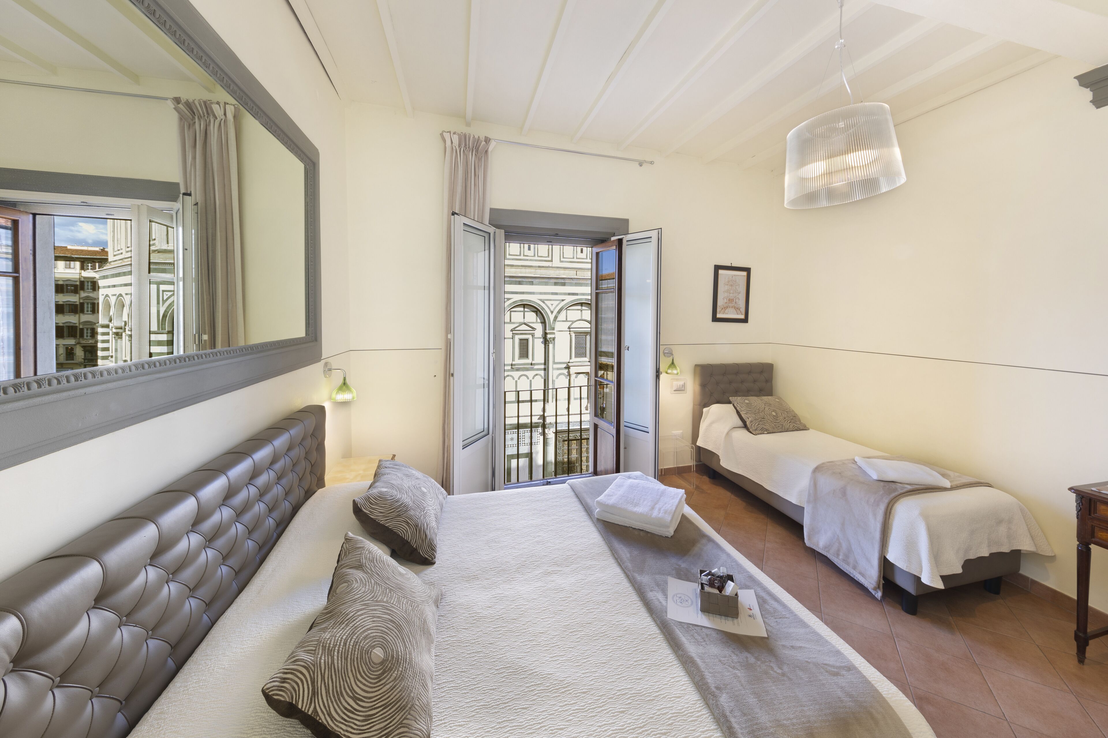 A hotel room with a large bed, a smaller bed, a sofa, a mirror, and a view of a building through an open door.