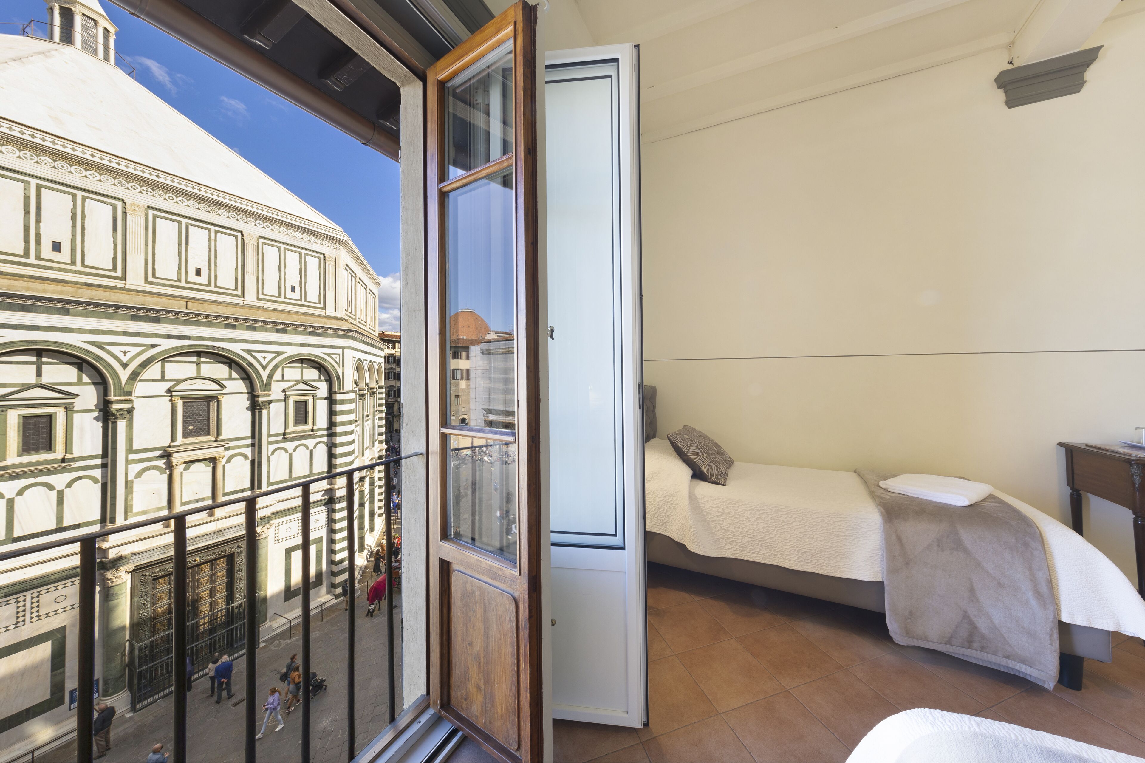 A room with a bed, a small bedside table, and a window overlooking a building with classical architecture.