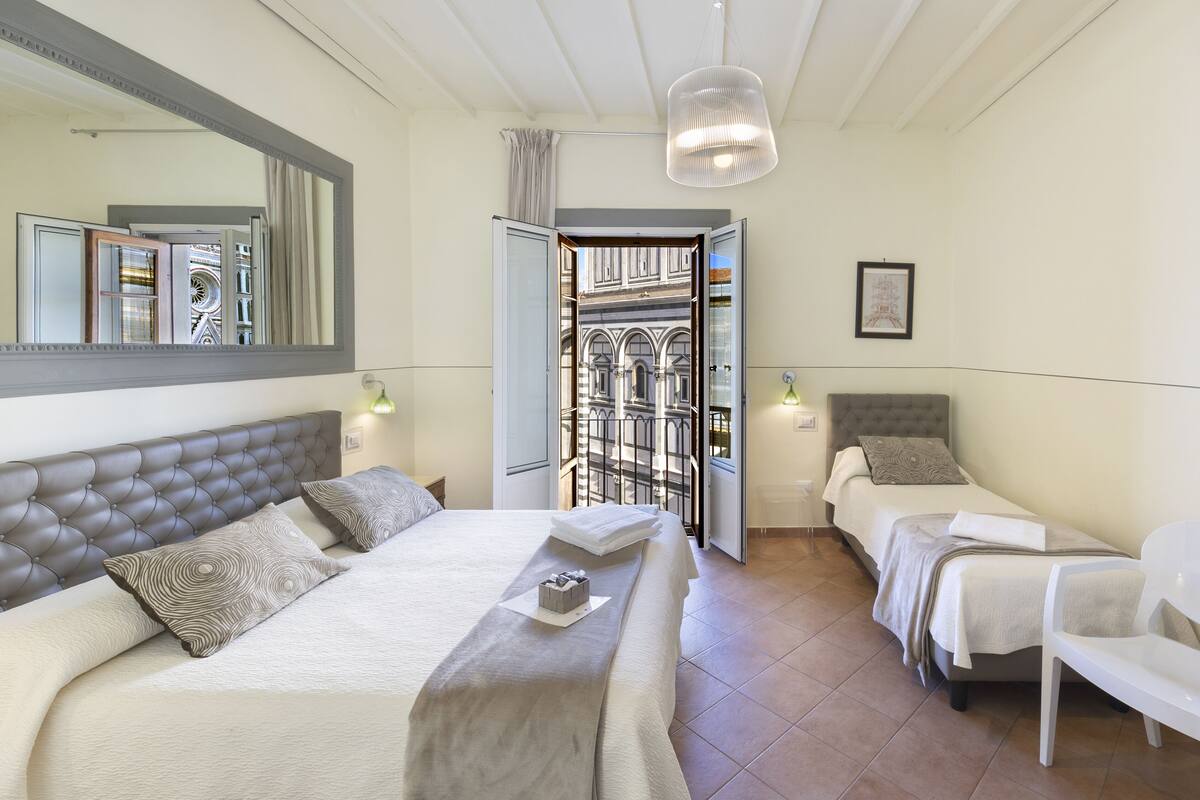 A hotel room with a large bed, two smaller beds, a sitting area, and a view of a building through an open door.