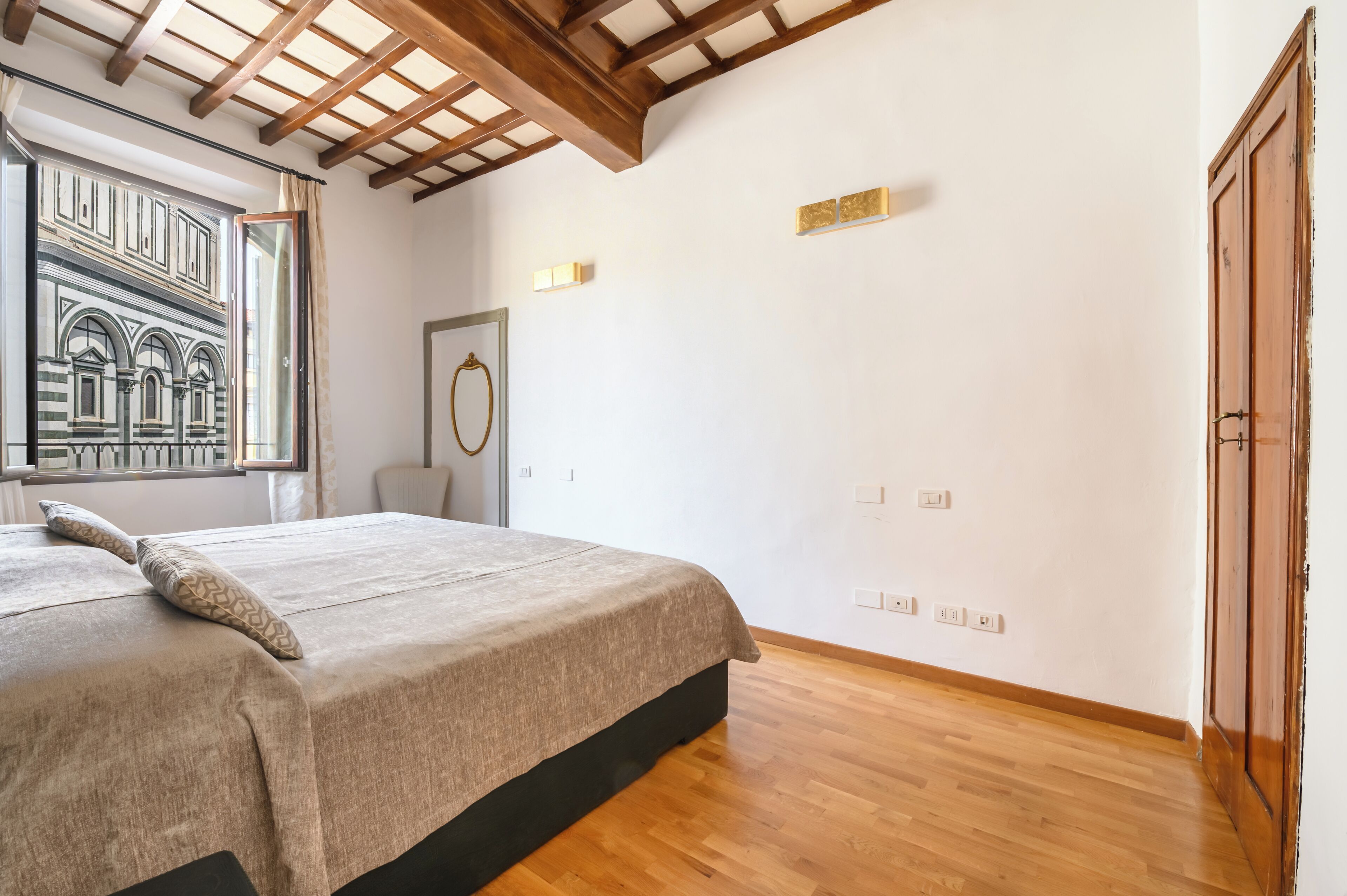 A bedroom with a bed, wooden floor, a window with a view of a building, and a door.