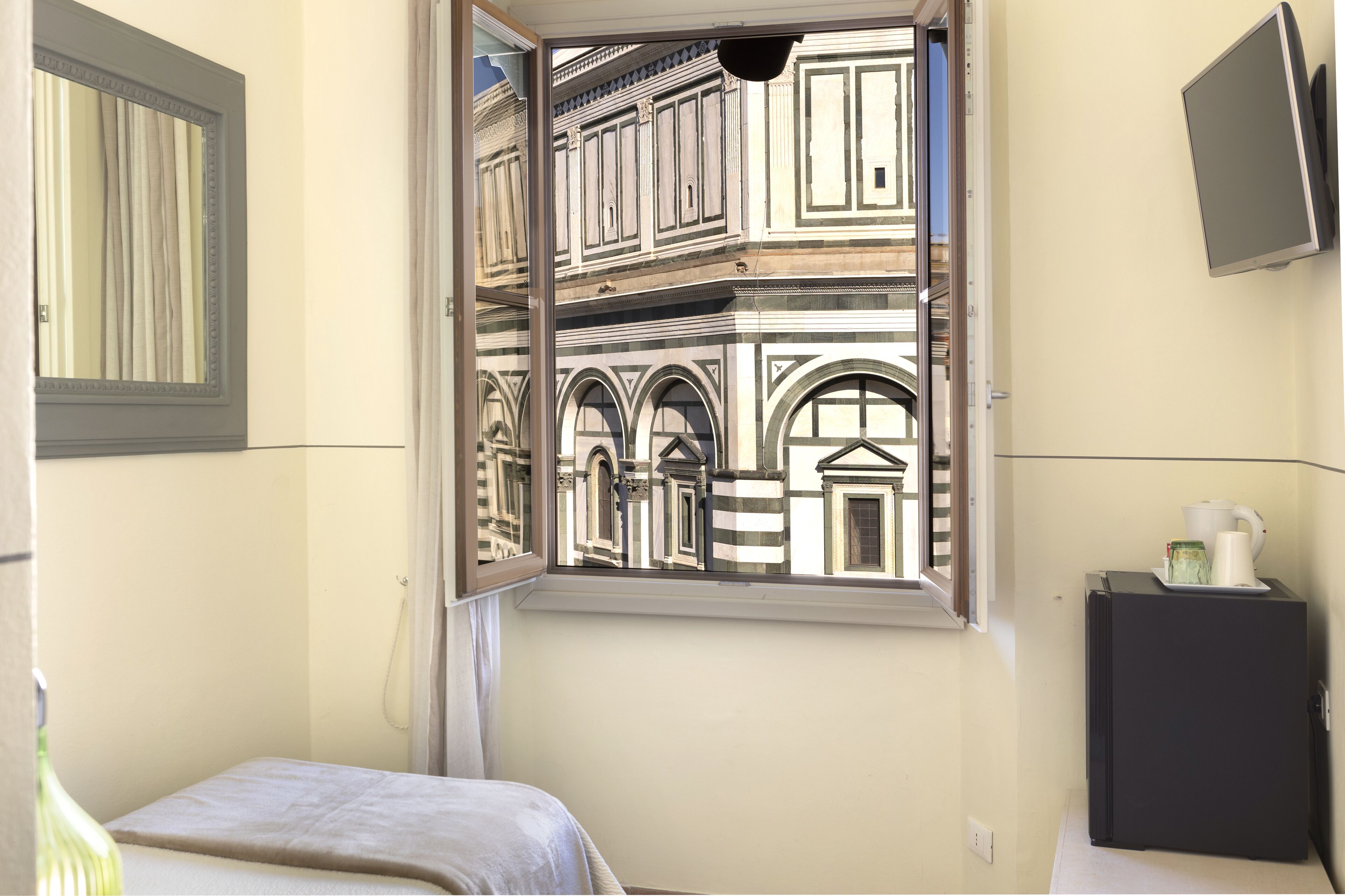 A room with a bed, a window with a view of a building, a television, and a small table with a cup and a container.