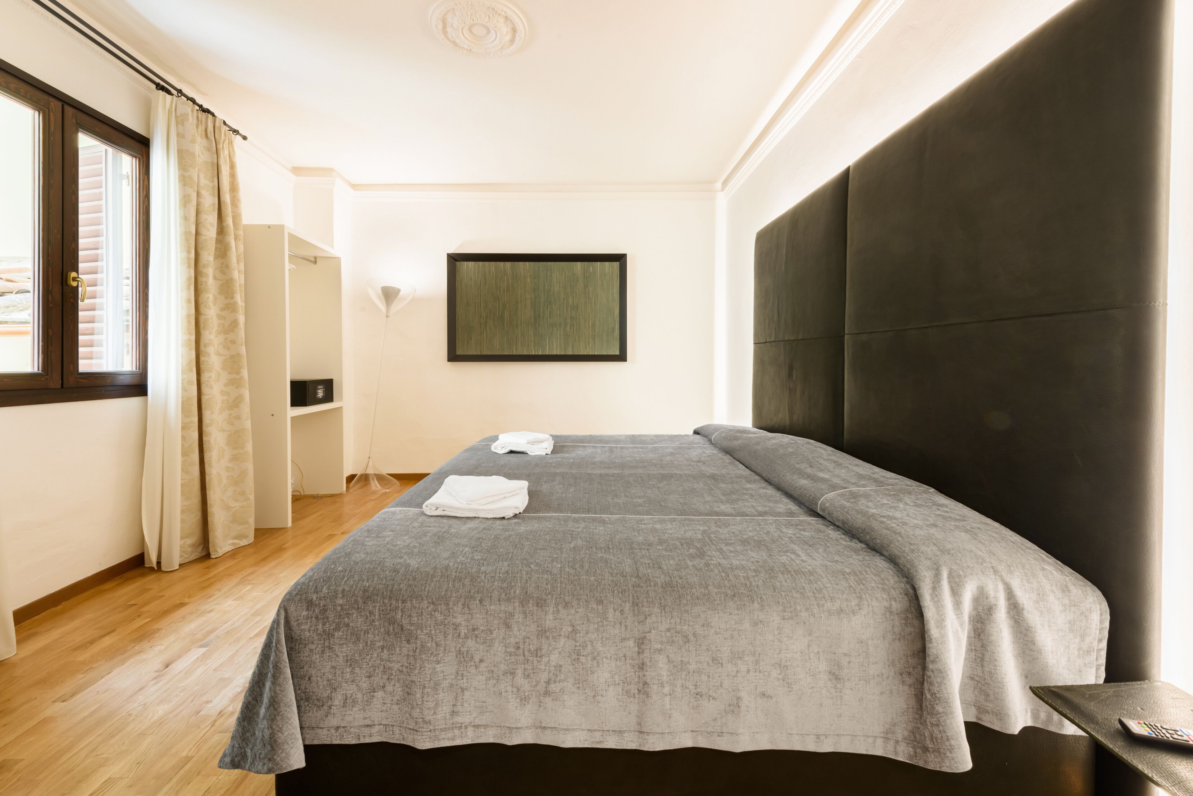 A modern hotel room with a large bed, a window with curtains, a wall-mounted TV, and a bedside table.
