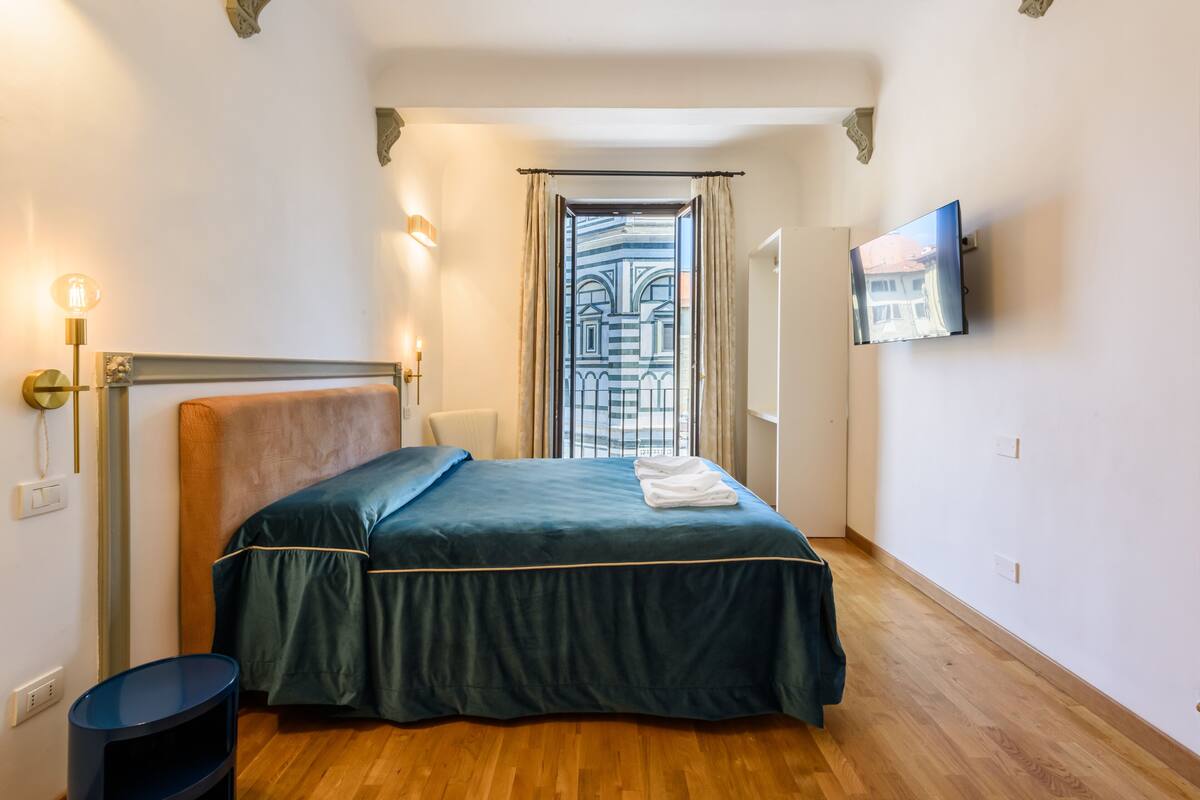 A hotel room with a bed, a TV mounted on the wall, a window with curtains, and a wooden floor.