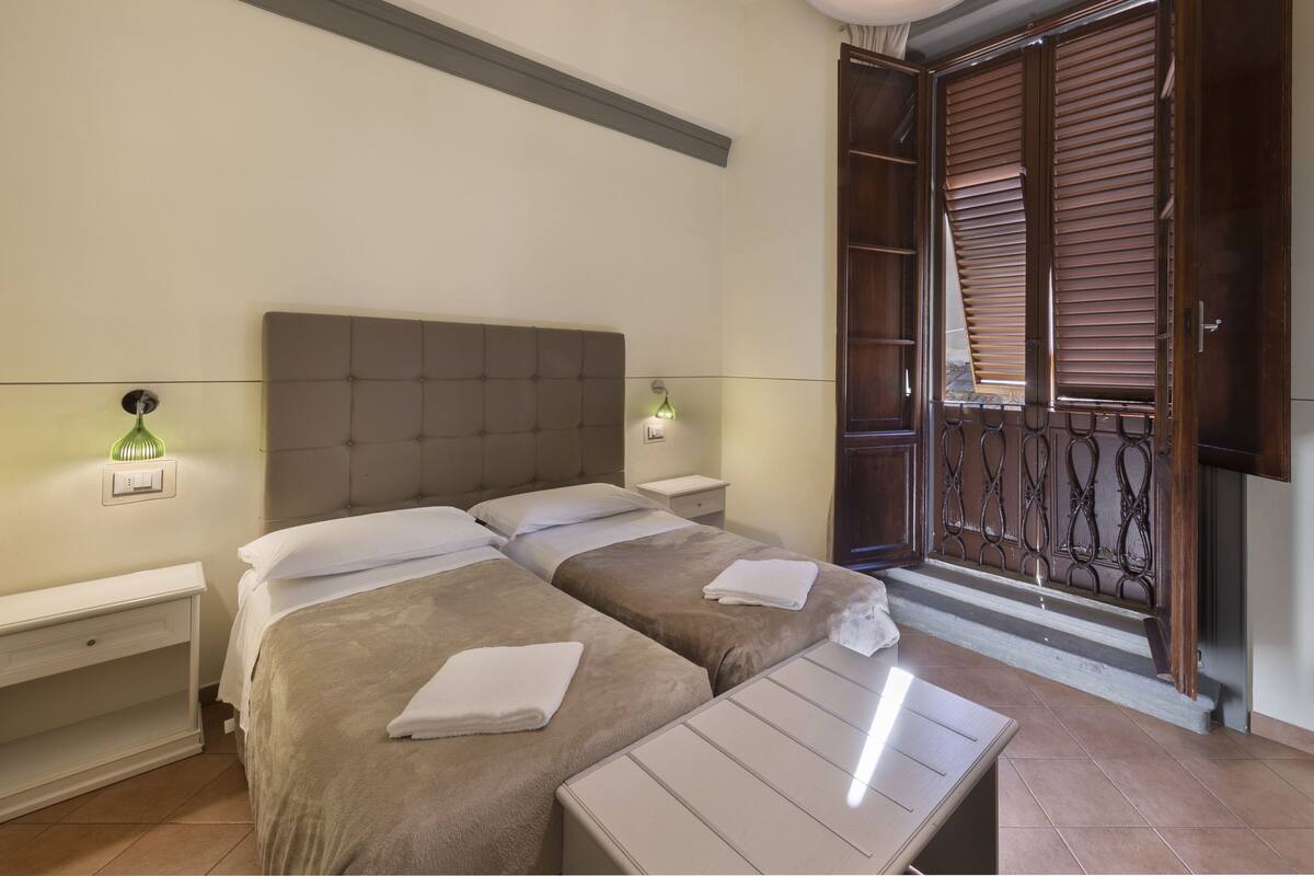 A hotel room with two beds, a small table, bedside lamps, and a balcony with wooden shutters.