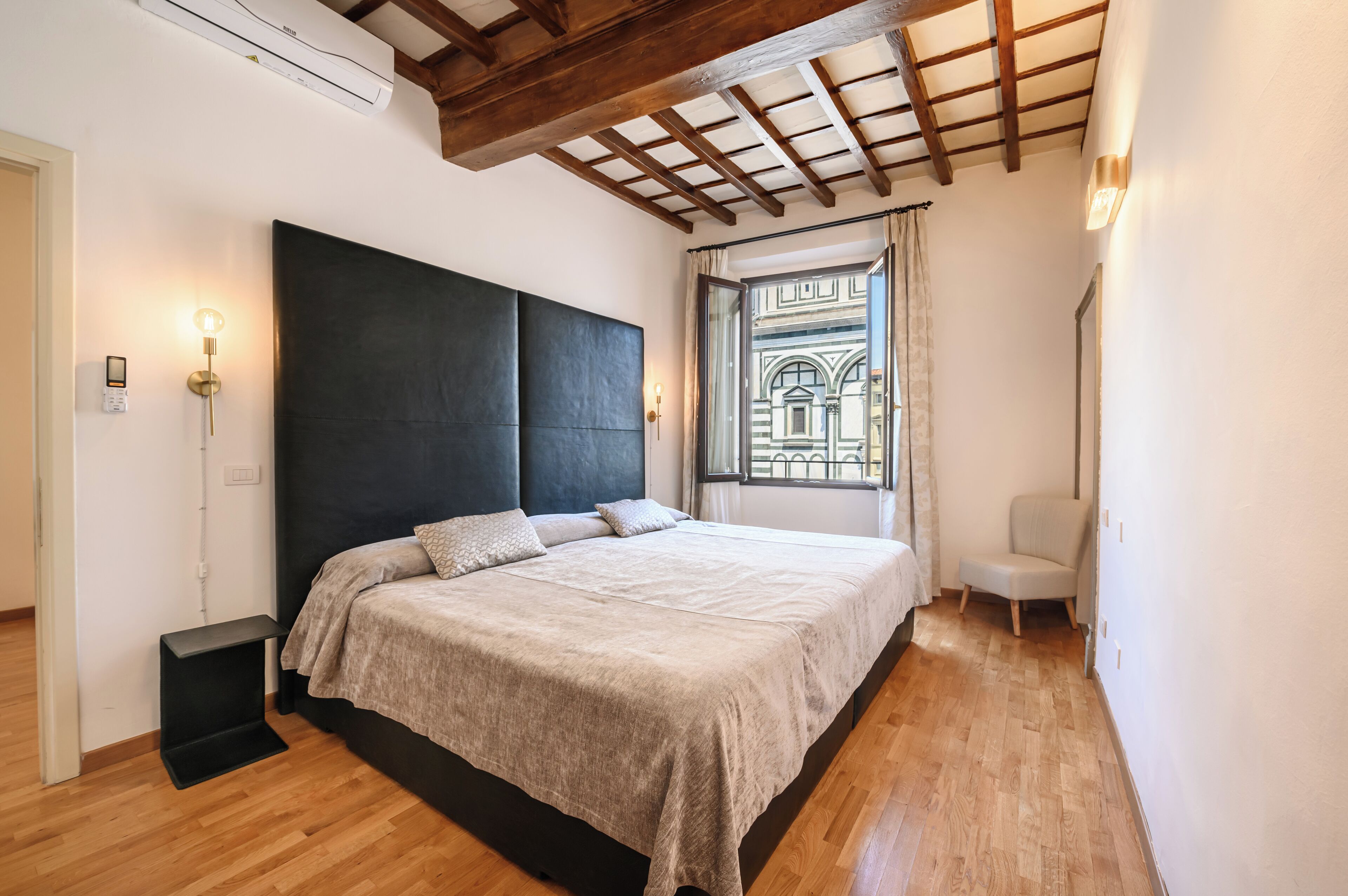 Un dormitorio con una cama grande, suelo de madera, cabecero oscuro, una ventana con vistas a un edificio y una silla.