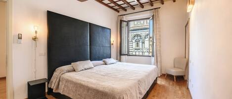 Un dormitorio con una cama grande, suelo de madera, cabecero oscuro, una ventana con vistas a un edificio y una silla.