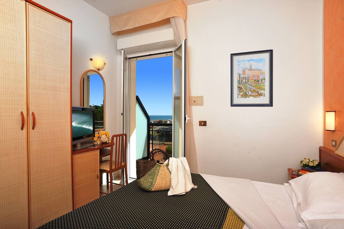 A hotel room with a bed, a desk with a computer, a chair, a wardrobe, and a balcony with a view.