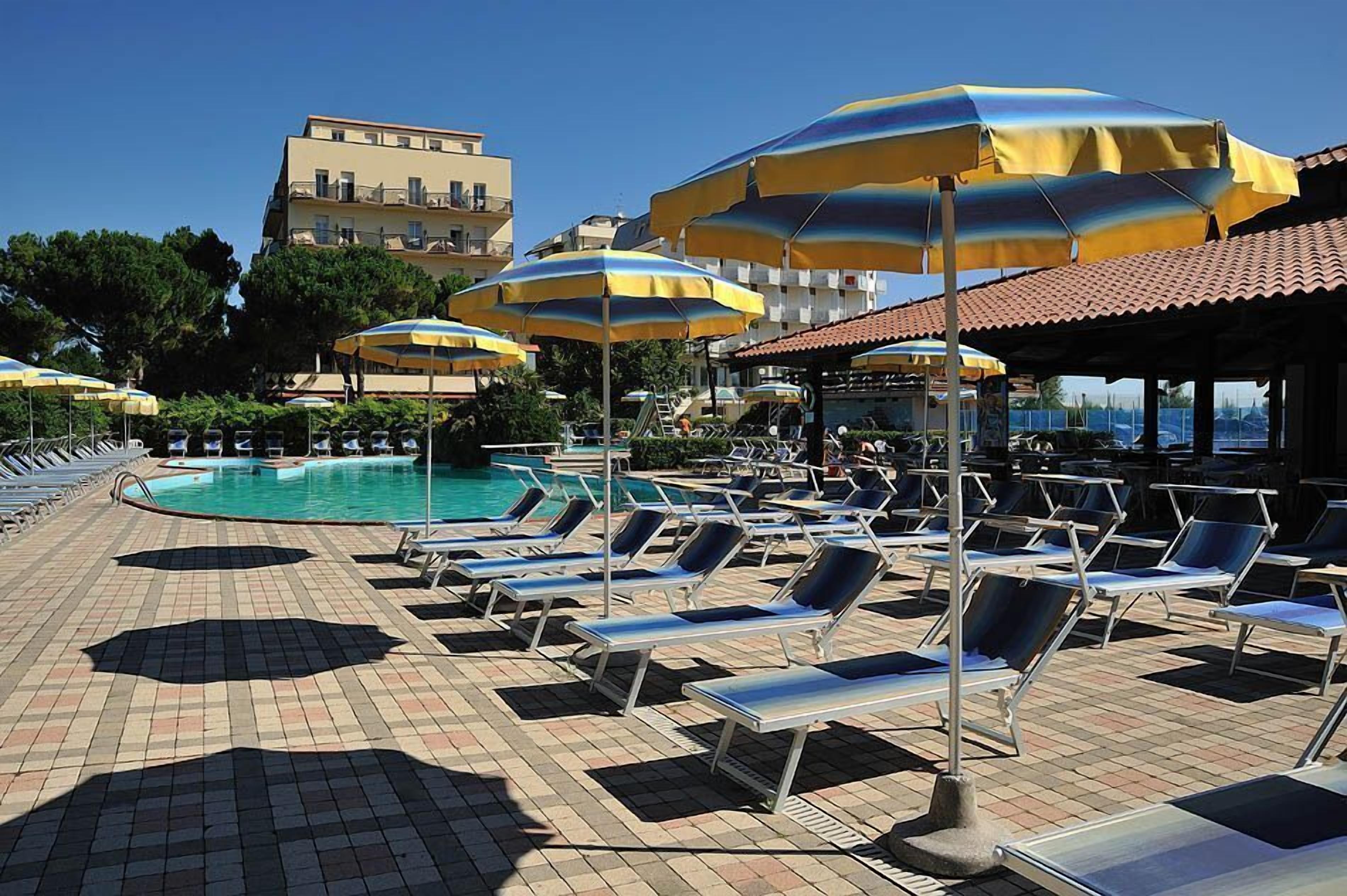 Zona junto a la piscina con tumbonas y sombrillas, una piscina azul cristalina y un edificio al fondo.