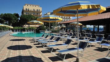 Zona junto a la piscina con tumbonas y sombrillas, una piscina azul cristalina y un edificio al fondo.