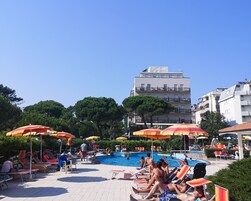 Area piscina con lettini e ombrelloni, sullo sfondo l'edificio di un hotel e un cielo azzurro intenso.